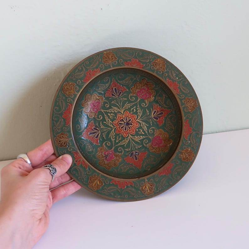May include: A round, decorative plate with a green background and floral design. The plate features red, pink, and gold accents. The plate is held by a hand with silver rings.