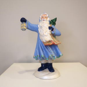 May include: A ceramic Santa Claus figurine in a light blue robe, holding a lantern and a small green Christmas tree. The figurine has a long white beard and is standing on a white base. The robe has a decorative trim.