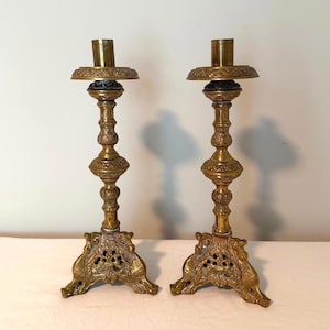 Puede incluir: Un par de candelabros de latón de estilo antiguo, altos, con bases texturizadas y ornamentadas con intrincados detalles recortados. Los tallos son delgados con nodos decorativos redondeados, y cada parte superior incluye un plato ancho.
