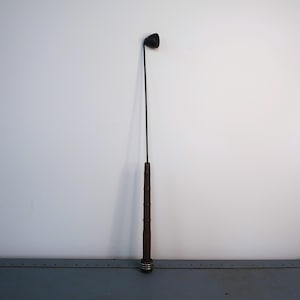 May include: A tall, antique-style candle snuffer. It features a black bell-shaped snuffer head, a long, slender black metal rod, and a brown, textured handle. The handle has silver-colored metal rings at the base.