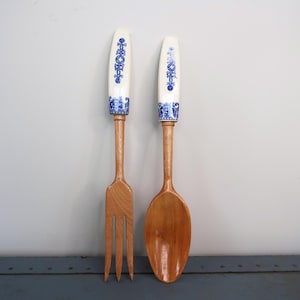May include: A wooden fork and spoon set. The fork has three tines, and the spoon has a rounded bowl. Both have light-colored wooden handles and white ceramic tops with blue floral designs. The set is likely for kitchen decor.