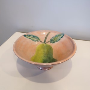 May include: A peach-colored ceramic bowl with a pedestal base. The interior features a hand-painted green pear with green leaves and a brown stem. The bowl is likely used for serving fruit or as a decorative piece.