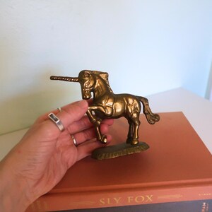 May include: A brass unicorn figurine with a detailed mane and tail. The mythical creature stands on a small base. The unicorn is a golden color and is displayed on top of a book.
