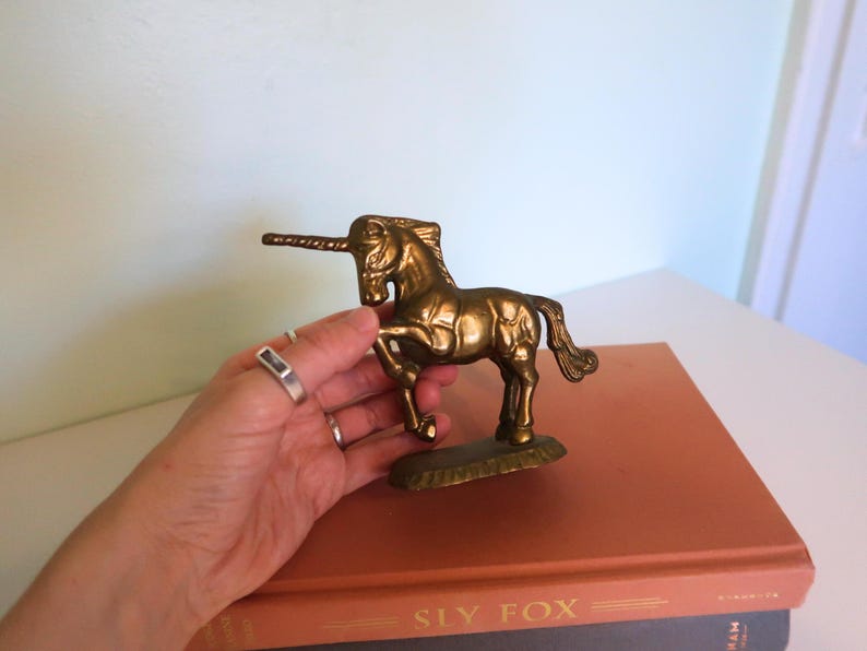 May include: A brass unicorn figurine with a detailed mane and tail. The mythical creature stands on a small base. The unicorn is a golden color and is displayed on top of a book.