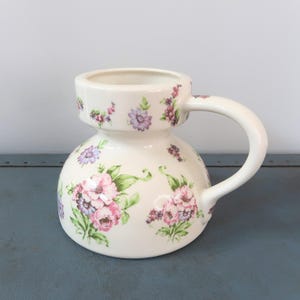 Puede incluir: Jarra de cerámica color crema con base bulbosa y parte superior cilíndrica. Decorada con un estampado floral de flores rosas, moradas y verdes. Tiene un asa curva. Ideal para servir bebidas.