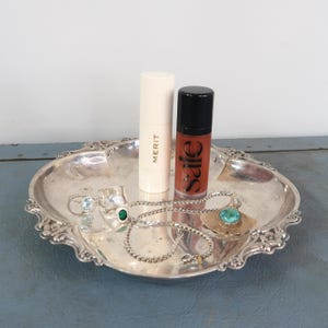Puede incluir: Una bandeja plateada ornamentada contiene productos de belleza y joyas. Incluye un tubo de lápiz labial, un brillo labial con texto "sale", anillos con piedras preciosas y un collar con un colgante turquesa. La bandeja tiene un borde decorativo festoneado.