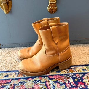 Könnte beinhalten: Ein Paar wadenhohe Stiefel aus beigem Leder mit abgerundeter Zehenpartie, einem gestapelten Blockabsatz und einem Schlupfdesign mit vertikalen Seitenlaschen. Die Stiefel haben sichtbare Nähte entlang des Rahmens und eine matte Oberfläche.