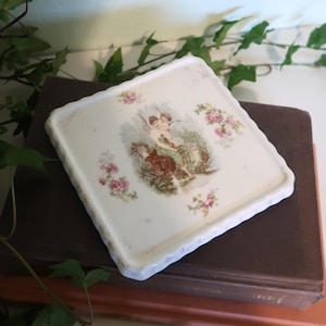 May include: Square, off-white ceramic tray with a scalloped edge. The tray features a central image of two cherubs and floral accents in pink and green. The tray is resting on a brown surface.