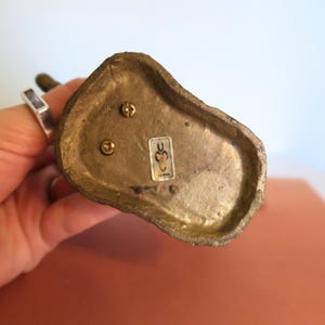 May include: Close-up of the bottom of a small, antique brass figurine. The base is irregularly shaped with three screws and a small sticker. The figurine is held by a person's hand, with a silver ring visible.