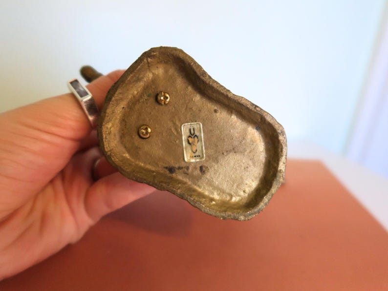 May include: Close-up of the bottom of a small, antique brass figurine. The base is irregularly shaped with three screws and a small sticker. The figurine is held by a person's hand, with a silver ring visible.
