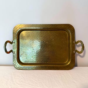 May include: A rectangular brass tray with rounded corners and two curved handles. The tray has a hammered texture and a slightly tarnished appearance, suggesting age. The handles are attached with small rivets.