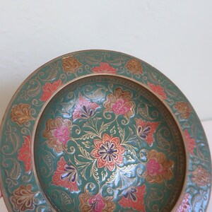 May include: A decorative, round, teal-colored plate with an intricate floral design. The plate features pink, red, and gold floral patterns. The rim is a gold color. The plate is likely made of metal.