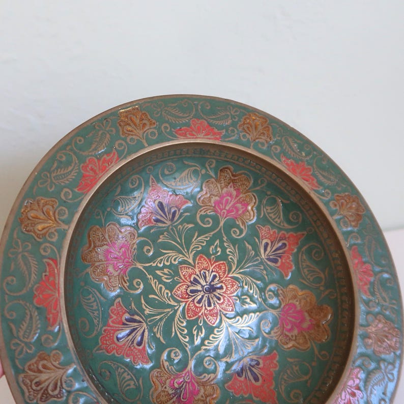 May include: A decorative, round, teal-colored plate with an intricate floral design. The plate features pink, red, and gold floral patterns. The rim is a gold color. The plate is likely made of metal.