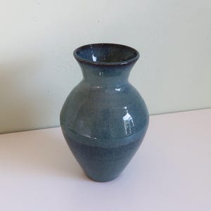 Puede incluir: Un jarrón de cerámica hecho a mano con un esmalte azul verdoso brillante. El jarrón tiene un cuerpo redondeado que se estrecha hacia una base estrecha y se ensancha en la parte superior. El borde es de un tono azul más oscuro. Este jarrón decorativo es adecuado para flores.