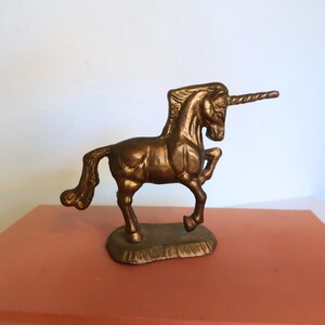 May include: A bronze-colored unicorn figurine with a spiraled horn and a detailed mane and tail. The mythical creature is posed on a small, textured base, and is displayed on a reddish-orange surface.