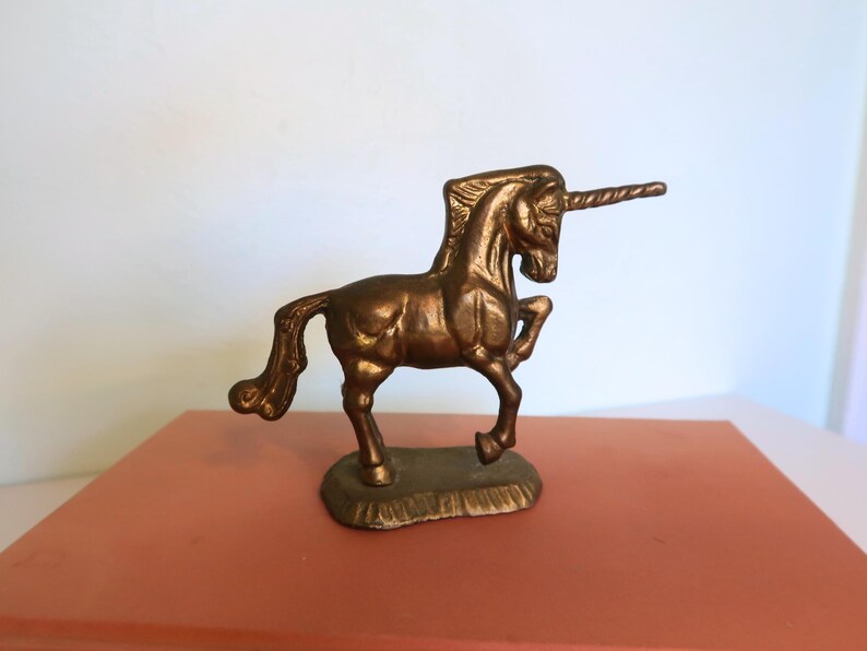 May include: A bronze-colored unicorn figurine with a spiraled horn and a detailed mane and tail. The mythical creature is posed on a small, textured base, and is displayed on a reddish-orange surface.