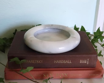 Pode incluir: Um cinzeiro de mármore branco com um design circular repousa sobre dois livros antigos. O cinzeiro tem uma superfície lisa e polida e uma bacia rasa. Os livros são castanhos escuros e cor de pêssego, com letras douradas.