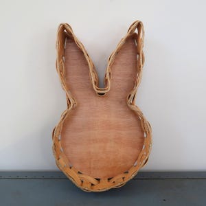 Puede incluir: Una cesta con forma de conejo con una base de madera marrón claro y laterales trenzados de color marrón claro. La cesta tiene orejas largas y erguidas y un cuerpo redondeado. Los lados trenzados están asegurados con pequeñas grapas plateadas.