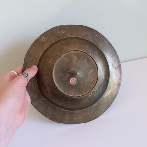 May include: A tarnished brass plate with a circular design. The plate has a raised rim and a central, smaller circular indentation. A small sticker on the plate reads "MADE IN INDIA".