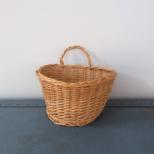 Pode incluir: Um cesto de vime tecido castanho claro, com formato curvo e alça trançada. O cesto está vazio e assenta numa superfície azul contra uma parede branca. Ideal para decoração.