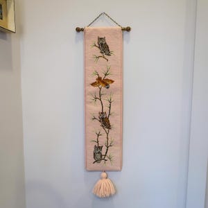 May include: A pink tapestry wall hanging featuring embroidered owls perched on branches. The artwork includes a brown leaf and a tassel at the bottom. The tapestry is suspended by a metal chain and wooden dowels.