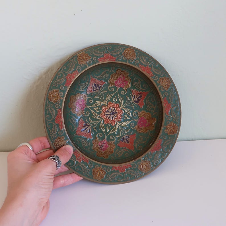 May include: A decorative, round plate with a green background and floral design. The plate features a central flower with red and orange petals, surrounded by pink and gold floral patterns. The rim has a gold border with more floral details.