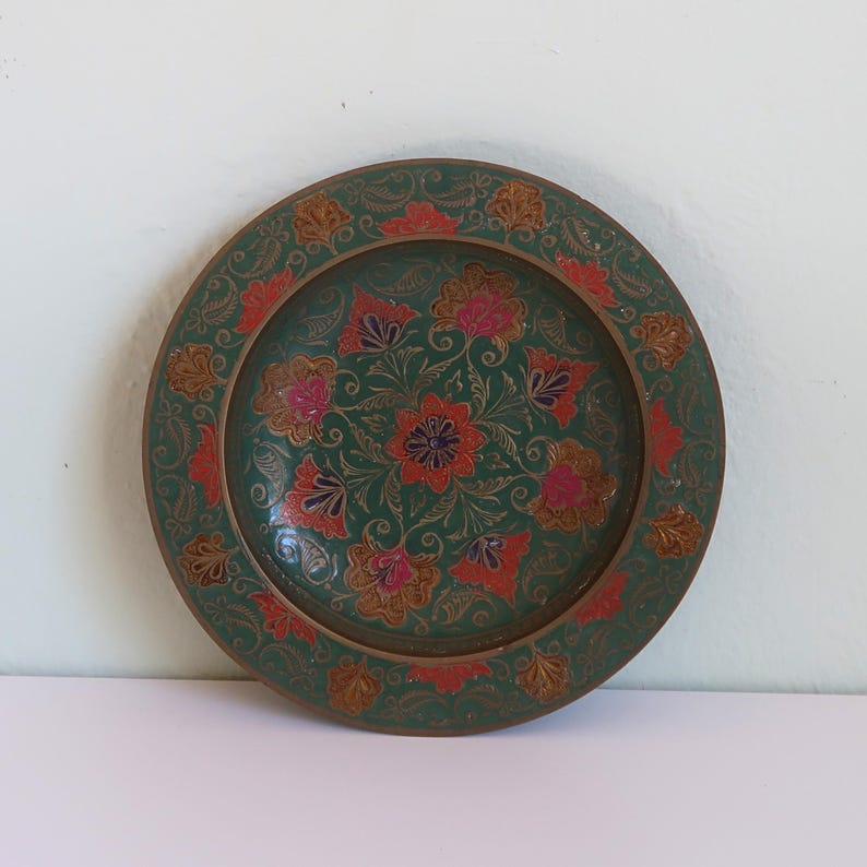 May include: A round, decorative plate with a green background and floral design. The plate features red, pink, and gold floral patterns. The rim has a gold border with a repeating floral motif. The plate is likely made of metal.