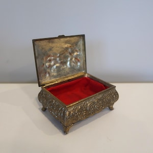 May include: An antique brass jewelry box with an open lid. The box has ornate floral detailing and a red velvet interior. The box is resting on four small feet.