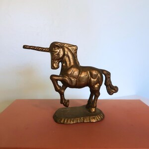 May include: A bronze unicorn figurine with a detailed mane, tail, and horn. The mythical creature is posed with one leg raised, standing on a textured base. The sculpture is displayed on a reddish-brown surface.