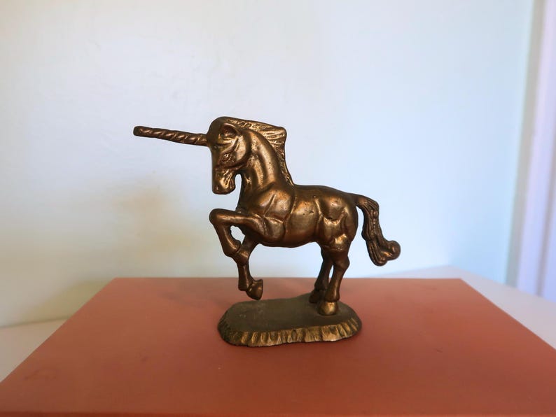 May include: A bronze unicorn figurine with a detailed mane, tail, and horn. The mythical creature is posed with one leg raised, standing on a textured base. The sculpture is displayed on a reddish-brown surface.