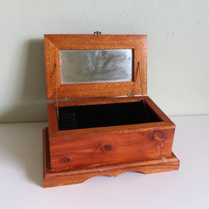 Peut inclure: Boîte à bijoux en bois ouverte, avec un couvercle à charnière et un petit miroir. La boîte est en bois brun rougeâtre et possède un intérieur en velours noir. Le couvercle est maintenu ouvert par de petites chaînes.
