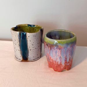 May include: Two ceramic cups with distinct glazes. The left cup is off-white with dark speckles and a vertical blue stripe. The right cup features a vibrant drip glaze pattern transitioning from a green rim to blue, purple, and coral pink tones.