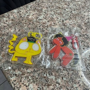 May include: Two acrylic keychains in clear plastic packaging. One is a yellow cat with a green hat and a cigarette, featuring a yellow clip. The other is a red, stylized figure with black hair and a pink cone hat, featuring a red clip.