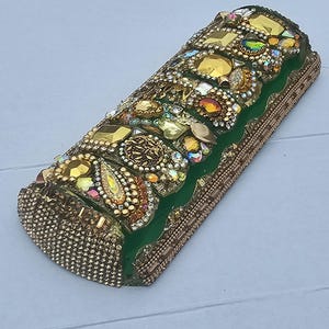 May include: A cylindrical, decorative object covered in gold-toned embellishments and gemstones. The top features a band of green fabric, adorned with various sizes and shapes of gold and iridescent jewels. The sides are covered in small, gold-colored beads.
