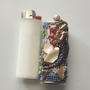 May include: A white lighter next to a decorative lighter. The decorative lighter features a mermaid design with red hair, a gold top, and a dark purple tail. The design is embellished with iridescent rhinestones, pearls, and shell accents.