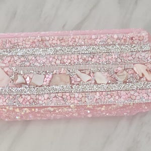 May include: A rectangular, pink, decorative box encrusted with small pink stones and silver rhinestones. The box is adorned with several seashells and has a shimmering, textured appearance. The overall design is elegant and eye-catching.