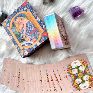 Pode incluir: Um baralho de tarot Wonderstruck com uma caixa ilustrada com uma figura loira em um vestido azul floral, um ma&ccedil;o com bordas hologr&aacute;ficas iridescentes e um leque de cartas com fundo rosa e um buqu&ecirc; de flores brancas, amarelas e azuis.