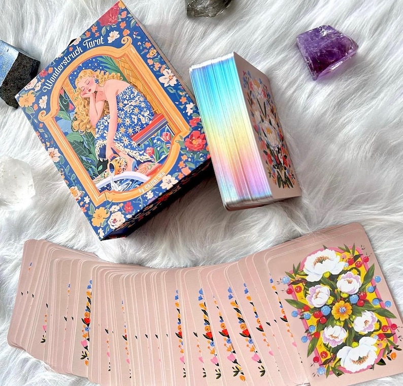 Pode incluir: Um baralho de tarot Wonderstruck com uma caixa ilustrada com uma figura loira em um vestido azul floral, um ma&ccedil;o com bordas hologr&aacute;ficas iridescentes e um leque de cartas com fundo rosa e um buqu&ecirc; de flores brancas, amarelas e azuis.