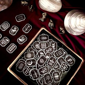Könnte beinhalten: Eine Sammlung kleiner, achteckiger schwarzer Plättchen mit weißen Strichzeichnungen von Tarot-inspirierten Symbolen. Viele Plättchen sind in einem quadratischen Tablett mit goldenem Rand angeordnet, während andere zusammen mit einem Würfel verstreut liegen.
