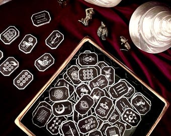 Pode incluir: Uma coleção de pequenas peças pretas octogonais com ilustrações de linhas brancas de símbolos inspirados no tarot. Muitas peças estão dispostas dentro de uma bandeja quadrada com borda dourada, enquanto outras estão espalhadas ao lado de um dado.