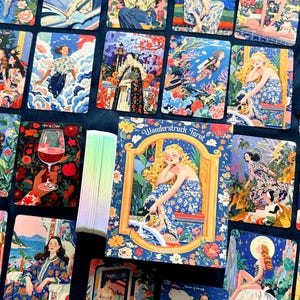 Pode incluir: Um baralho de cartas de tarot Wonderstruck com ilustra&ccedil;&otilde;es coloridas e detalhadas de figuras em v&aacute;rios cen&aacute;rios. A caixa exibe uma figura loira com um vestido floral azul, e as bordas das cartas mostram um gradiente de cores pastel.