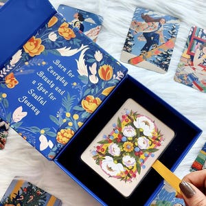 Pode incluir: Uma caixa azul com um padr&atilde;o floral e o texto Born for Everyday Beauty and a Love for Soulful Journey. Dentro da caixa h&aacute; um baralho com uma ilustra&ccedil;&atilde;o floral no verso. V&aacute;rias outras cartas ilustradas est&atilde;o espalhadas por perto.