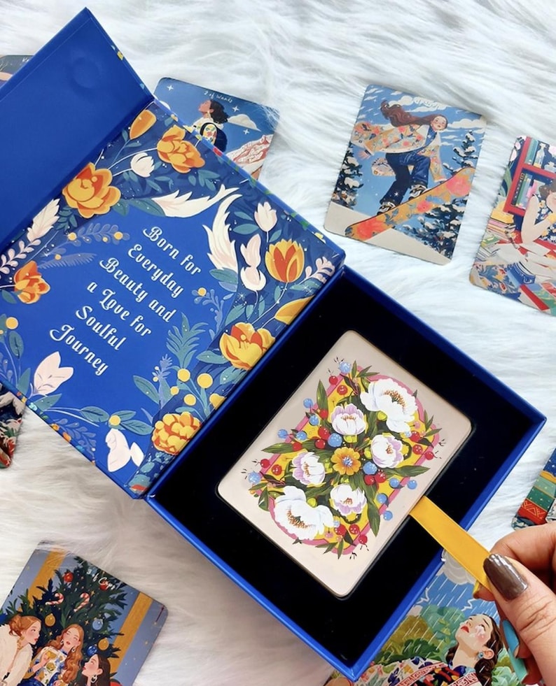 Pode incluir: Uma caixa azul com um padr&atilde;o floral e o texto Born for Everyday Beauty and a Love for Soulful Journey. Dentro da caixa h&aacute; um baralho com uma ilustra&ccedil;&atilde;o floral no verso. V&aacute;rias outras cartas ilustradas est&atilde;o espalhadas por perto.