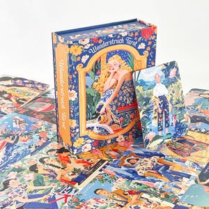 Puede incluir: Una baraja de tarot Wonderstruck con caja y cartas que presentan ilustraciones coloridas y detalladas de figuras en diversos entornos. La caja azul muestra una imagen central de una persona rubia con un vestido floral y el texto Wonderstruck Tarot.
