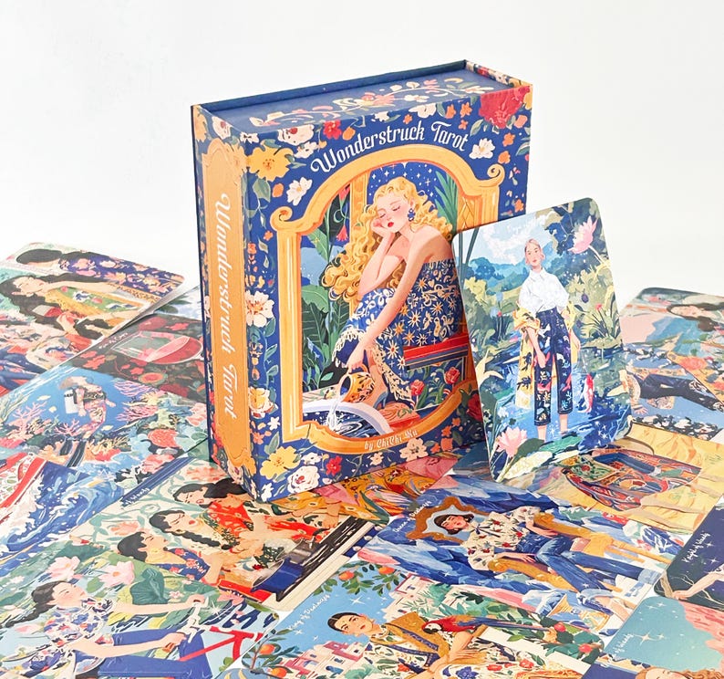 Pode incluir: Um baralho de tarot Wonderstruck com caixa e cartas apresentando ilustra&ccedil;&otilde;es coloridas e detalhadas de figuras em v&aacute;rios cen&aacute;rios. A caixa azul exibe uma imagem central de uma pessoa loira em um vestido floral com o texto Wonderstruck Tarot.