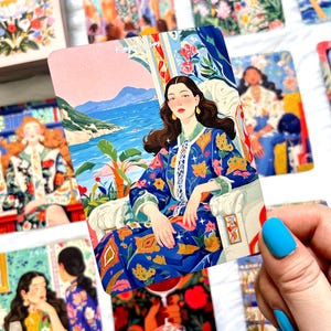 Pode incluir: Uma carta de tarot retangular intitulada King of Cups com a ilustra&ccedil;&atilde;o de uma pessoa de cabelos longos e escuros vestindo um manto azul estampado, sentada numa cadeira branca perante uma paisagem costeira com &aacute;gua azul, montanhas e plantas.