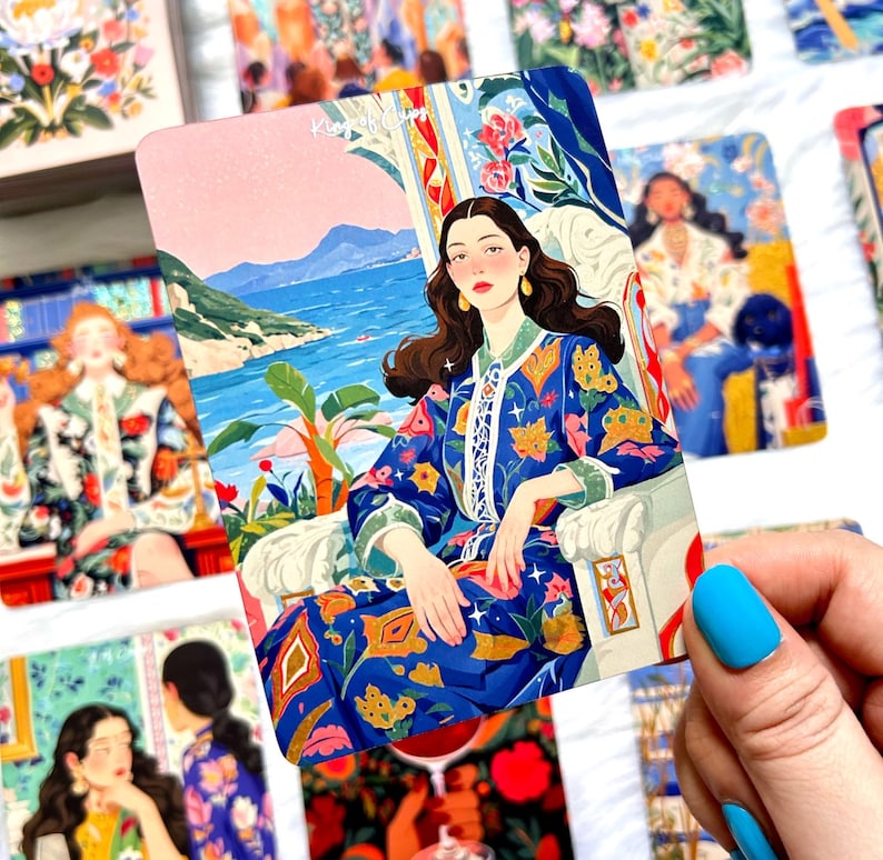 Pode incluir: Uma carta de tarot retangular intitulada King of Cups com a ilustra&ccedil;&atilde;o de uma pessoa de cabelos longos e escuros vestindo um manto azul estampado, sentada numa cadeira branca perante uma paisagem costeira com &aacute;gua azul, montanhas e plantas.