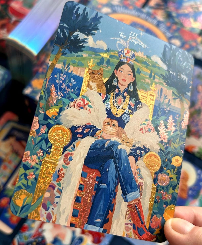 Pode incluir: Uma carta de tarot retangular intitulada The Empress III com a ilustra&ccedil;&atilde;o de uma pessoa usando uma coroa, roupas azuis estampadas e uma estola de pele branca, sentada num trono dourado com dois gatos, rodeada por flores e vegeta&ccedil;&atilde;o.