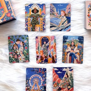 Pode incluir: Uma cole&ccedil;&atilde;o de sete cartas de tarot retangulares com ilustra&ccedil;&otilde;es coloridas e detalhadas de figuras e cenas, incluindo The Lovers, The Chariot e The World, dispostas numa superf&iacute;cie branca ao lado de uma caixa ilustrada a condizer.