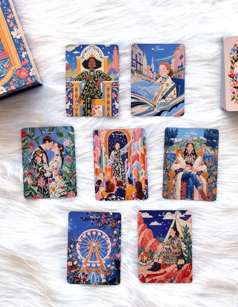 Pode incluir: Uma cole&ccedil;&atilde;o de sete cartas de tarot retangulares com ilustra&ccedil;&otilde;es coloridas e detalhadas de figuras e cenas, incluindo The Lovers, The Chariot e The World, dispostas numa superf&iacute;cie branca ao lado de uma caixa ilustrada a condizer.